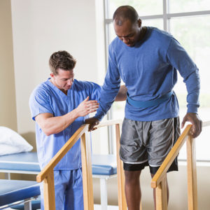 A man is scrubs is helping another man walk in physical therapy.