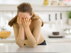 A woman is leaning on her kitchen counter with her hands on her face.