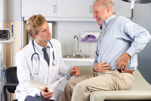 An older man is sitting on the table in the dotor's office pointing to his left side as the doctor looks.