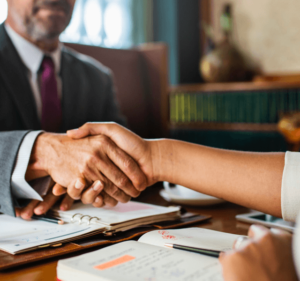 A man in a tie is shaking someone's hand across his desk.