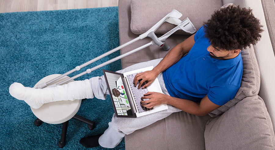 An aerial view of a man on a video call with his doctor as his foot is propped up in a cast.