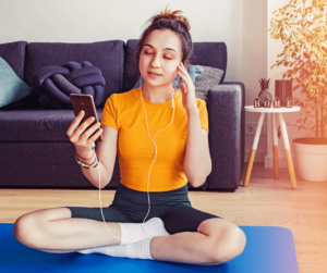 Young woman sitting on a yoga mat with headphones in listening while holding her phone.