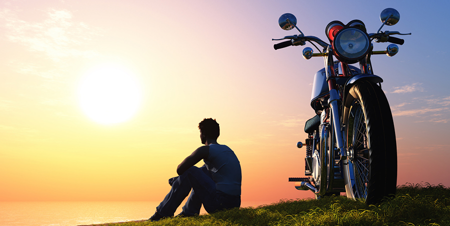 A guy on a hillside is watching the sunset next to his motorcycle.
