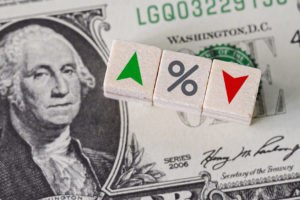 Small wood block of a green up arrow, percent sign, and red down arrow on a one dollar bill.